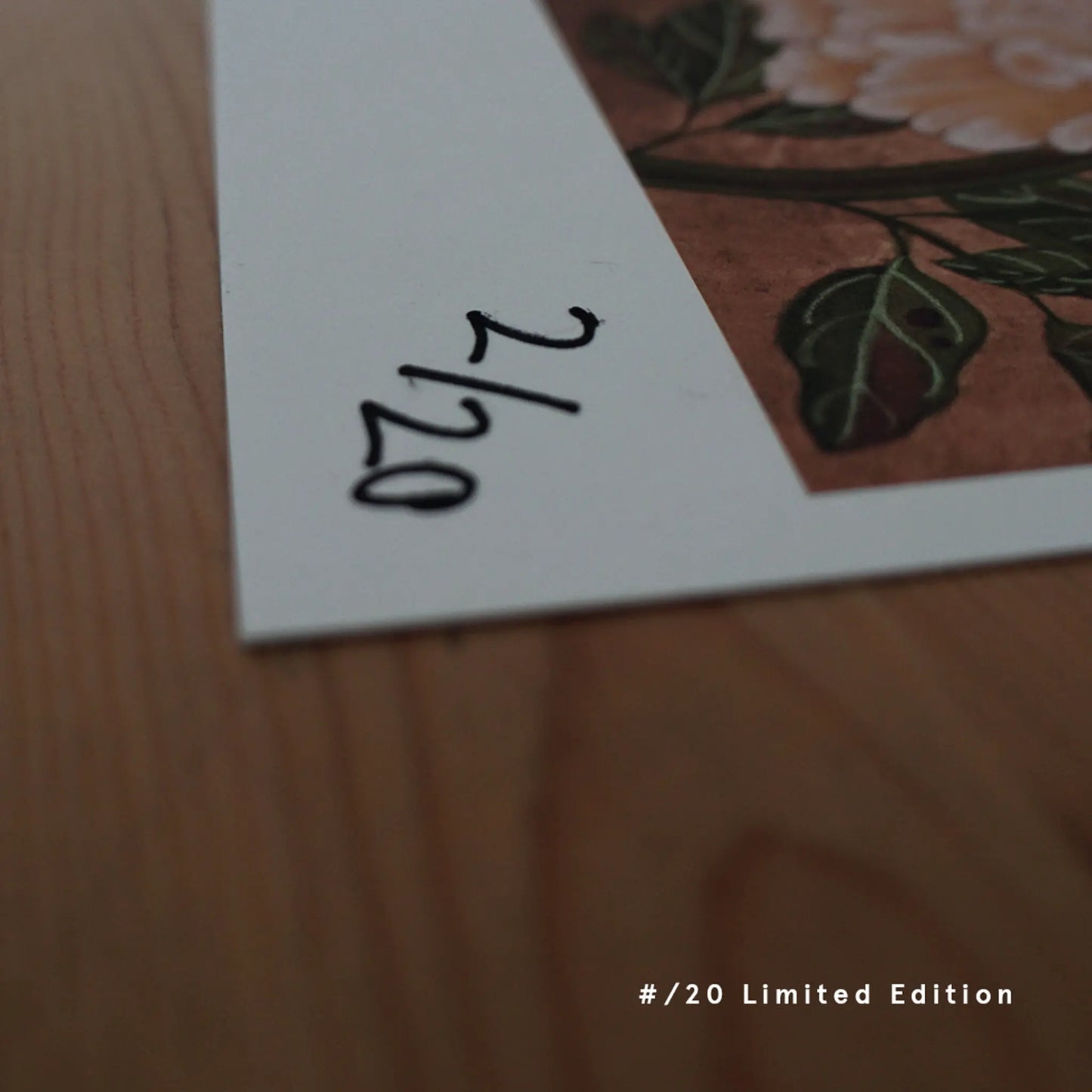 A close-up of a hand-numbered "2/20" limited-edition fine art print of "love" by Ryuku Otsuka. Resting on a warm wooden table, the print edge showcases a detailed pink and white peony with green leaves, offering a quiet, authentic sanctuary of traditional craft for collectors.