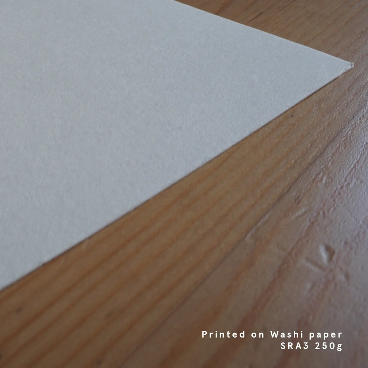 A close-up photograph showing the corner of a sheet of off-white Japanese textured paper resting on a warm wooden table surface, featuring text in the lower right corner reading "Printed on Washi paper SRA3 250g."