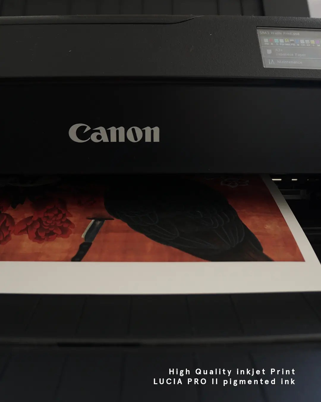 A close-up view of a fine art print emerging from a Canon ImagePROGRAF printer display panel. The artwork being printed depicts a dark bird silhouette, red floral elements, and a knife against an warm orange-toned background, with white text overlay at the bottom right reading "High Quality inkjet Print LUCIA PRO II pigmented ink."
