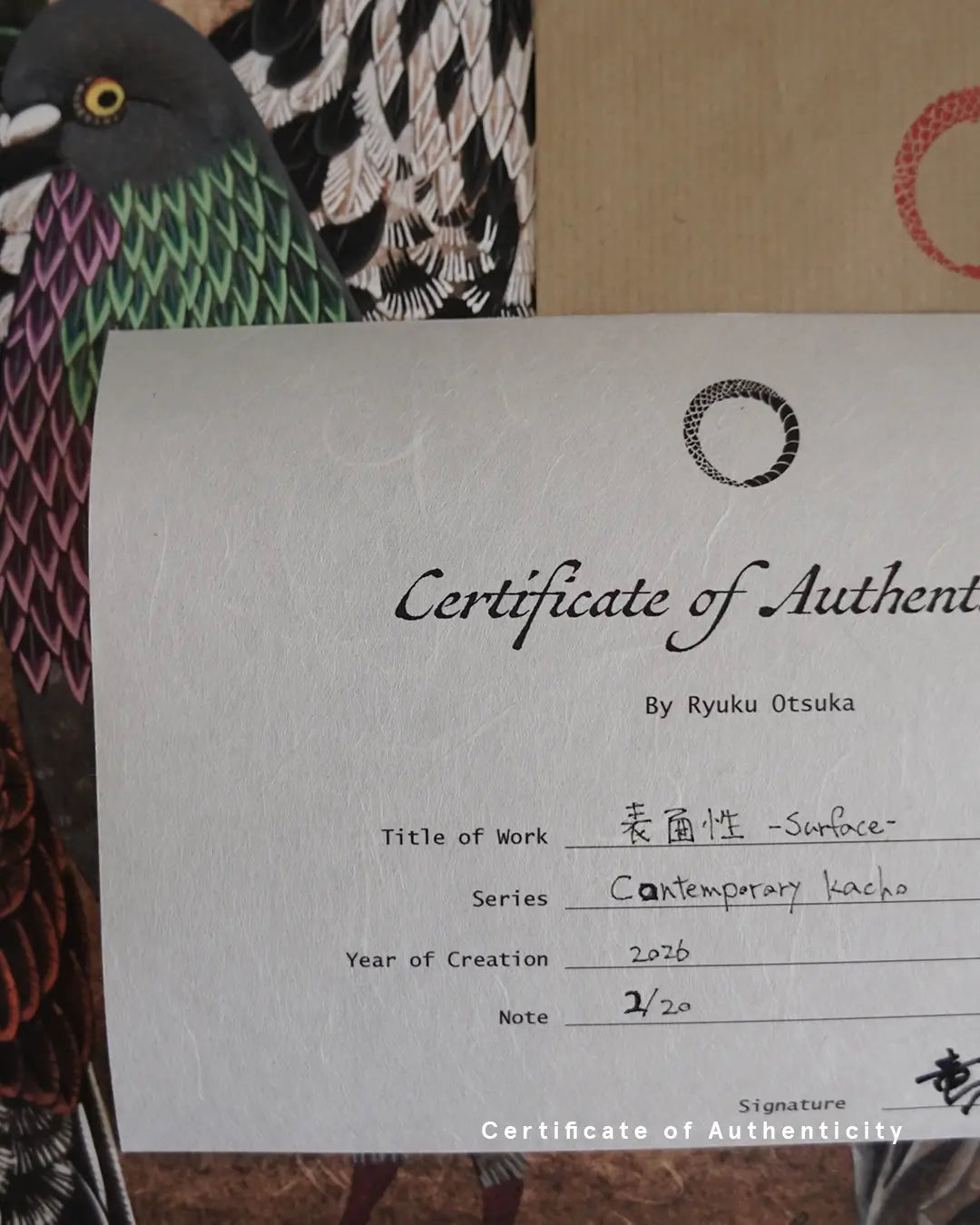 A close-up photograph of a Certificate of Authenticity printed on textured white washi paper for the fine art print "表面性 - Surface -" by Ryuku Otsuka. The document lists the series "Contemporary kacho," creation year 2026, limited edition numbering "2/20," and the artist's signature in Japanese kanji at the bottom right, set against an artwork featuring an urban pigeon with iridescence-patterned feathers.