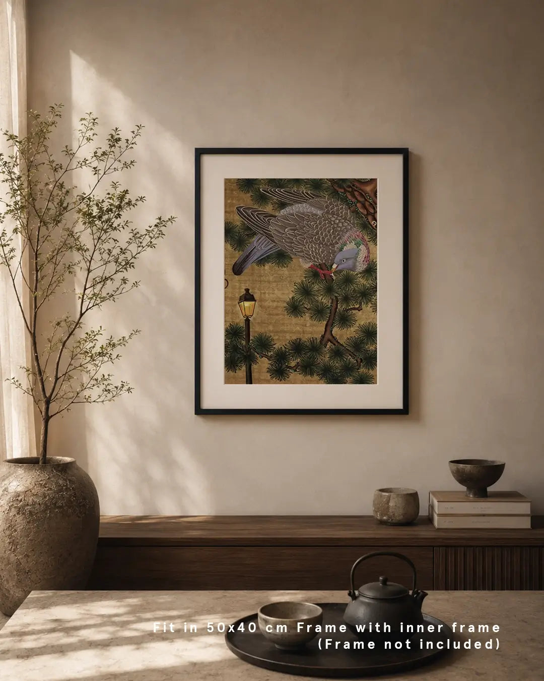 An interior display mockup showing the framed artwork "郷愁 - Nostalgia -" hanging on a neutral wall above a wooden console table. The fine art print features a detailed wood pigeon perched on pine branches near a lit street lamp against a textured gold background. The room setting includes a potted indoor tree, ceramic vessels, books, and a tea set, with text overlay reading "Fit in 50x40 cm Frame with inner frame (Frame not included)."