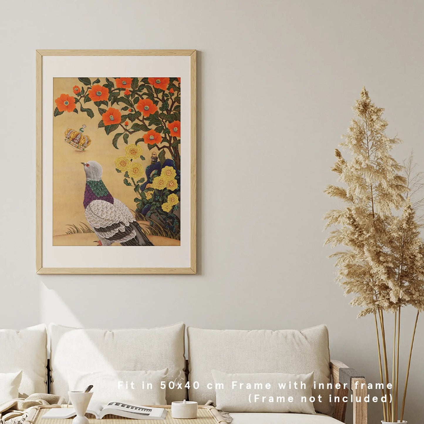 A framed fine art print by Ryuku Otsuka hanging on a neutral, off-white wall in a bright, modern living room setting. The print—featuring a detailed pigeon gazing up at a floating royal crown surrounded by orange blossoms and yellow flowers on a warm, textured gold background—is set within a light wood frame. Placed above a comfortable cream-colored sofa with plush pillows and alongside tall dried pampas grass, the scene demonstrates how the print fits a 50x40 cm frame to elevate slow-living home decor.