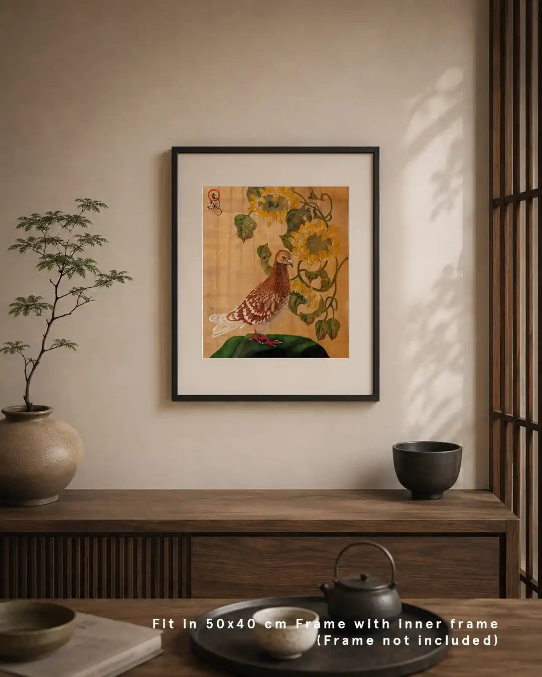 An interior mockup displaying a framed Japanese Nihonga art print hanging on a warm neutral wall above a dark wooden sideboard. The artwork shows a detailed brown and white pigeon standing alongside vibrant yellow sunflowers on a textured gold background. The serene room features a potted bonsai, a ceramic teaset on a tray, and text at the bottom reading "Fit in 50x40 cm Frame with inner frame (Frame not included)."