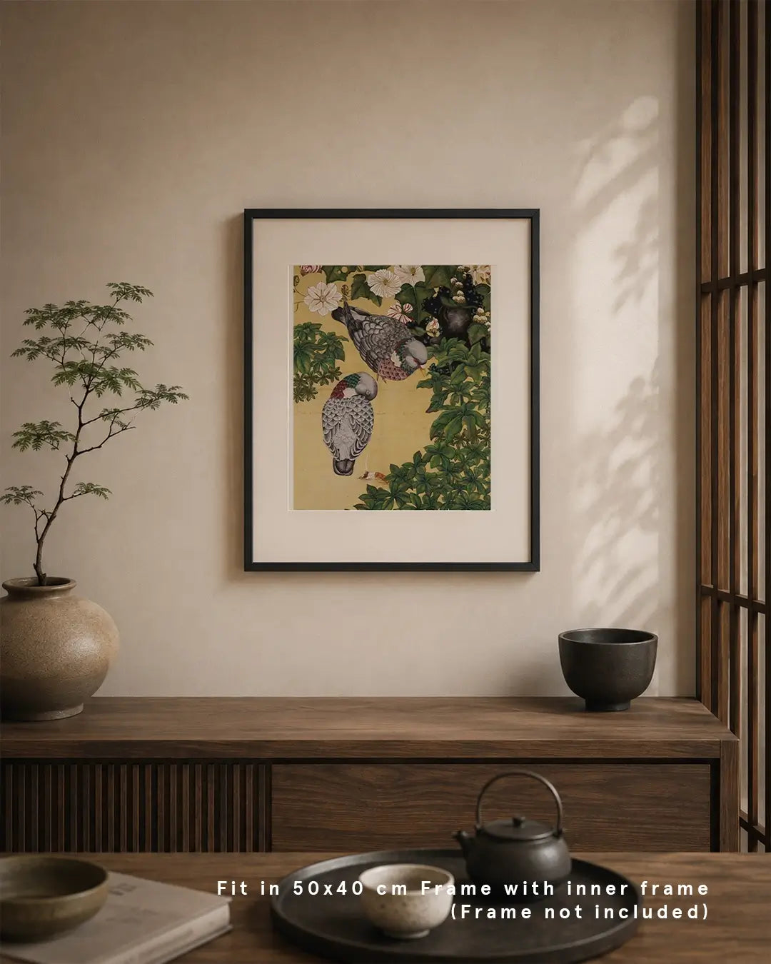 An interior wall mockup displaying a framed Japanese Nihonga art print mounted on a warm beige wall. The artwork depicts two urban pigeons amid leafy branches and delicate white blossoms on a golden background. The frame is positioned above a dark wood sideboard with a potted plant, ceramic tea set, and text overlay at the bottom reading "Fit in 50x40 cm Frame with inner frame (Frame not included)."