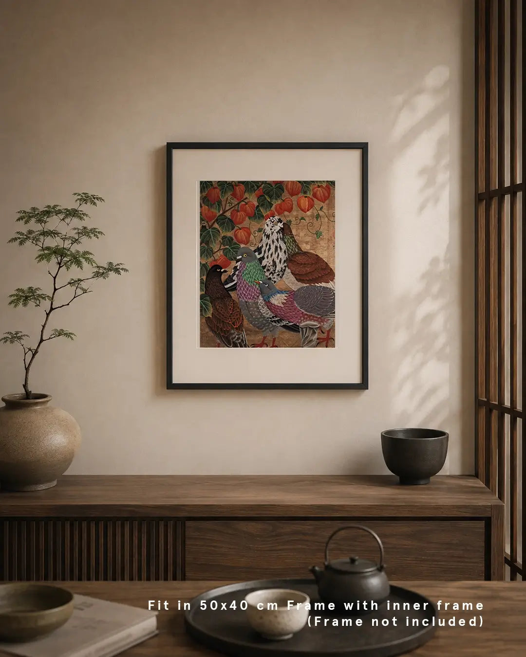 An interior mockup displaying a framed Japanese Nihonga art print mounted on a warm beige wall above a dark wood console table. The artwork depicts four urban pigeons gathered under orange Chinese lantern plant lanterns on a textured gold background. The serene room setting features a potted plant, ceramic tea set, and text overlay at the bottom reading "Fit in 50x40 cm Frame with inner frame (Frame not included)."