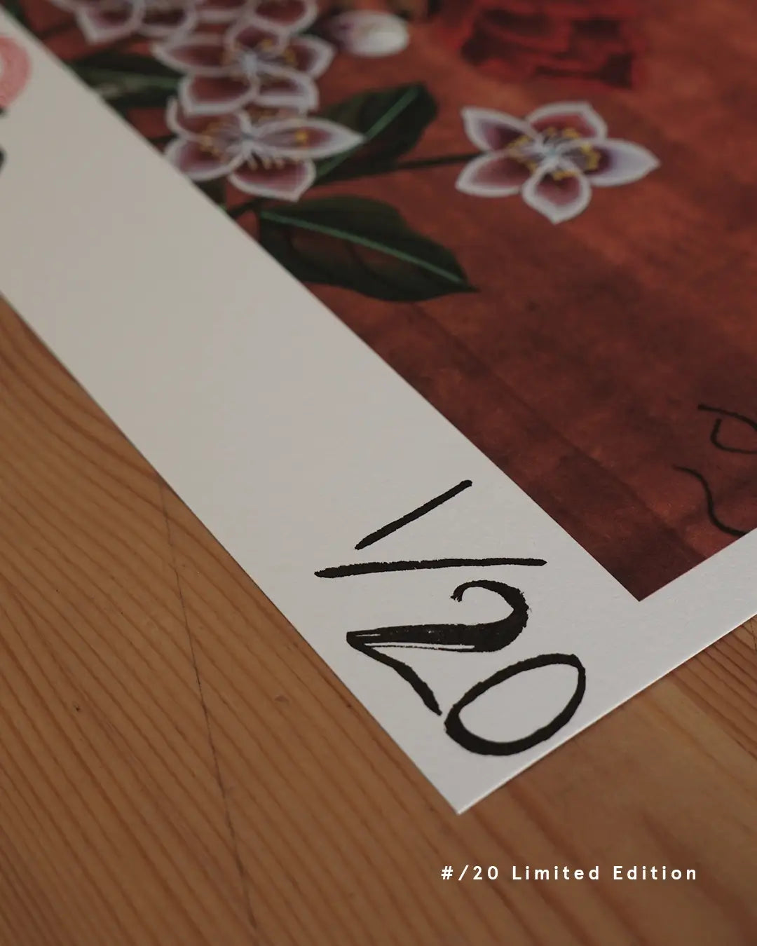 A close-up detail shot of the bottom-left corner of a fine art print on a wooden surface, displaying hand-written edition numbering "1/20" on the white margin. The artwork above features white plum blossom flowers with subtle pink hues against a rich reddish-brown background, with clean text overlay in the bottom right corner reading "#/20 Limited Edition."
