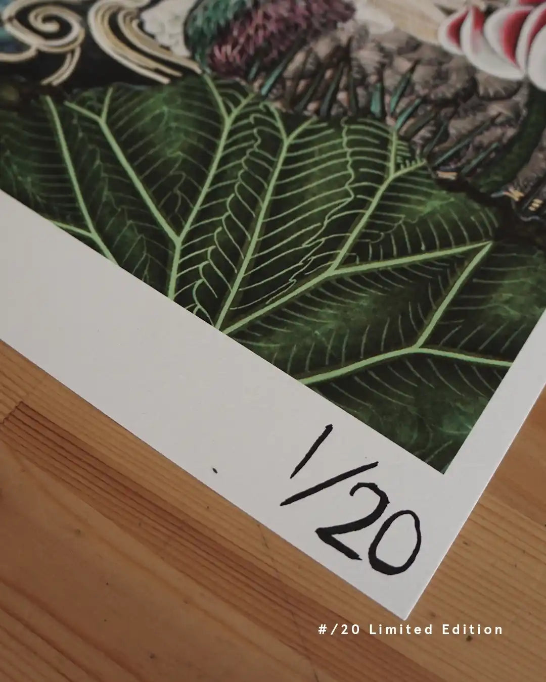 An angled close-up view showing the hand-numbered edition mark "1/20" on the white margin of a fine art print resting on a light wooden table, with rich green botanical leaf illustrations featured above and text overlay reading "#/20 Limited Edition."