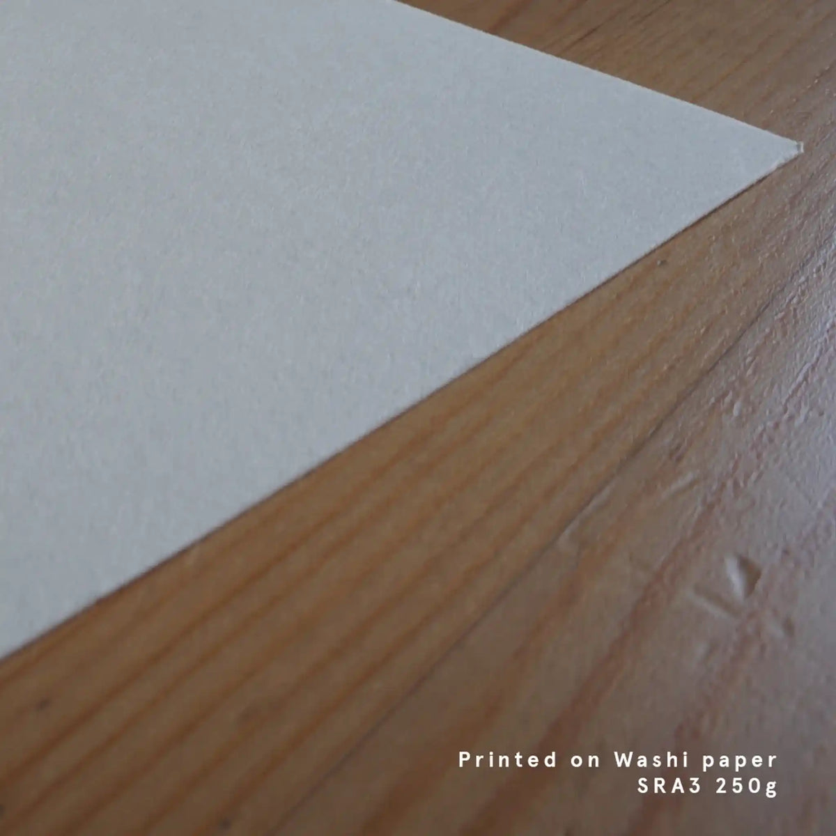 A detailed close-up shot showing the corner of a textured, off-white sheet of Japanese Washi paper resting on a warm wood desk, featuring white text overlay in the bottom right corner reading "Printed on Washi paper SRA3 250g."