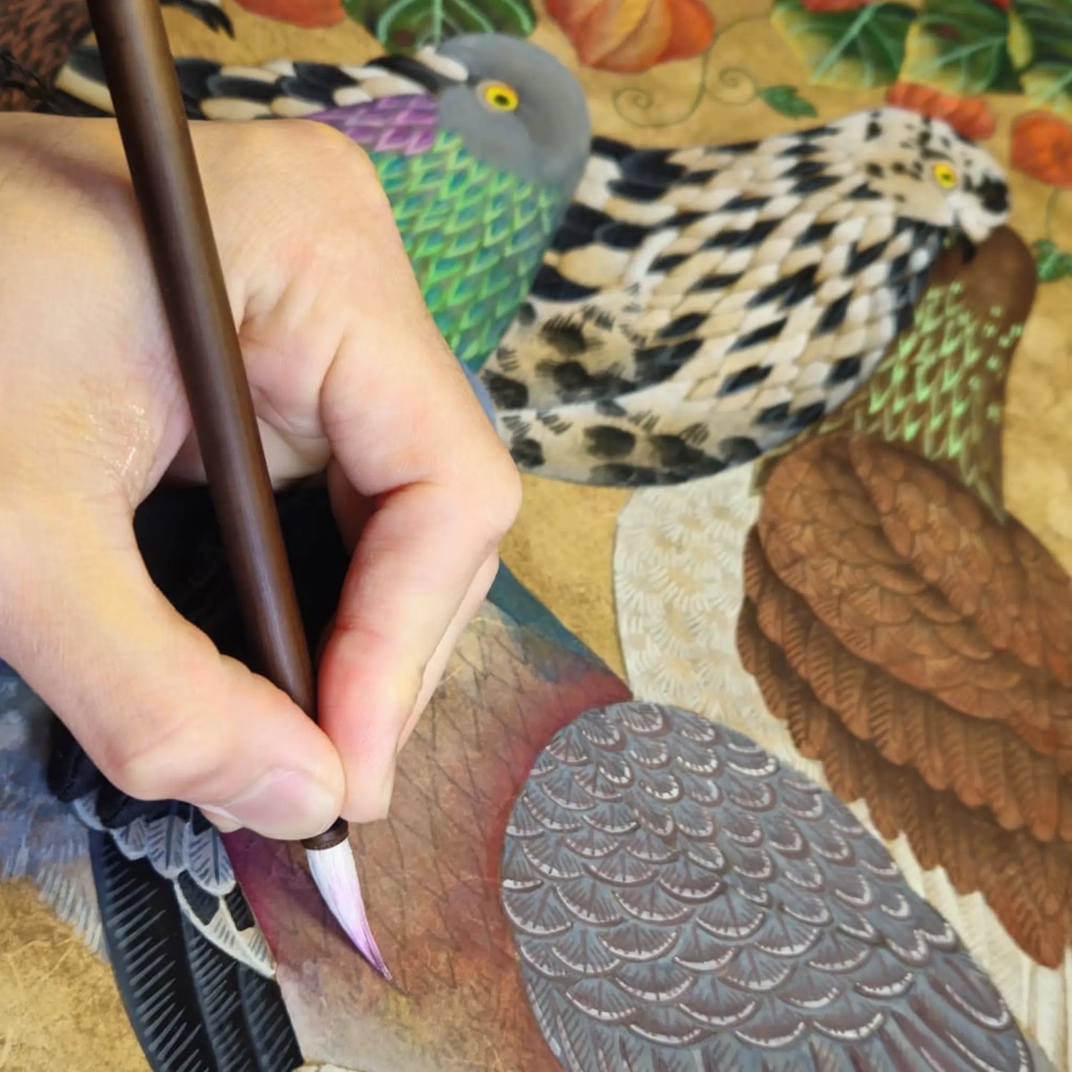 A close-up of Ryuku Otsuka's hand using a traditional wooden brush to paint detailed pigeon feathers onto textured paper. The image captures the delicate, patient brushwork of the Nihonga technique, with several colorful, stylized pigeons resting on a warm, organic-toned background, illustrating a slow-living commitment to craft.